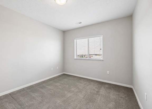 Bright apartment bedroom with carpeted flooring, neutral walls, and a window with blinds providing natural light.