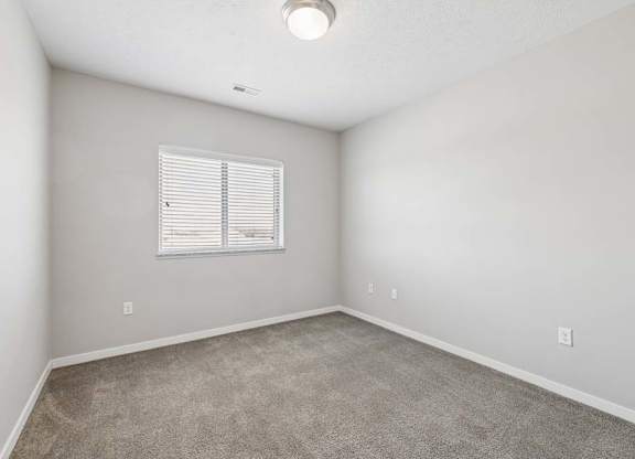 Bright apartment bedroom with carpeted flooring, neutral walls, and a window with blinds that provides natural light.