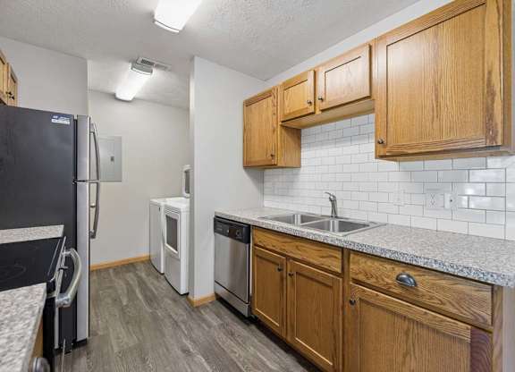 Galley-style apartment kitchen with wood cabinetry, a subway tile backsplash, stainless steel appliances, and an adjacent in-home laundry room.