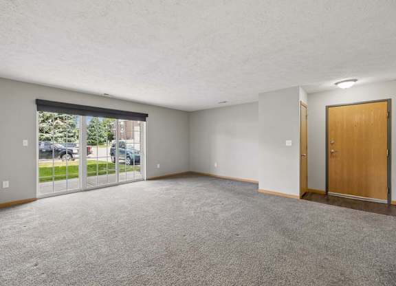 Spacious apartment living room with carpeted flooring, a large sliding glass door, and an open entryway with natural light.