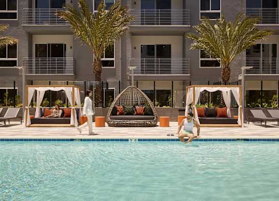 A woman is sitting on the edge of a pool in front of a building with palm trees.