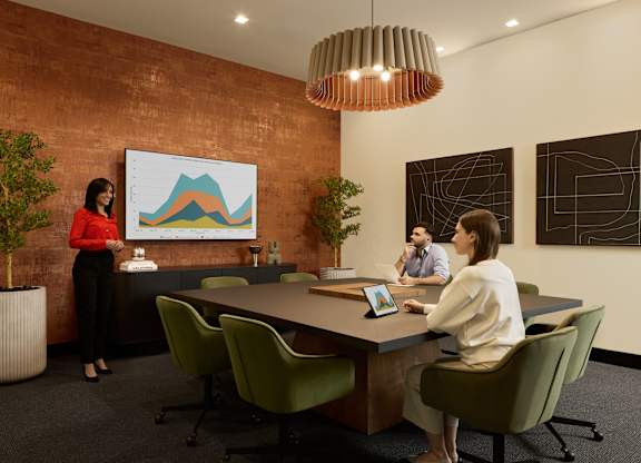 A woman in a red top stands in front of a whiteboard with a graph on it, while two other people sit at a table with laptops.