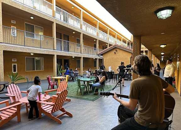A man is playing guitar in a courtyard with people sitting around a table.