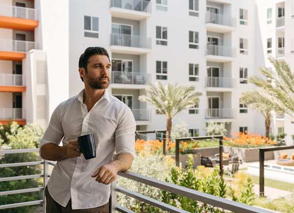 A man in a white shirt is standing on a balcony with a black mug in his hand.