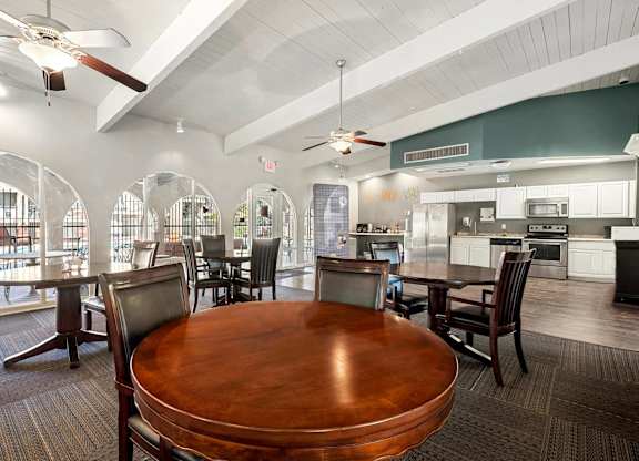 A large wooden table is surrounded by chairs in a spacious room with a kitchen in the background.