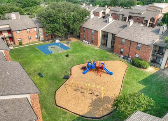 A playground with a slide, swings, and a basketball hoop in a grassy area surrounded by brick buildings.