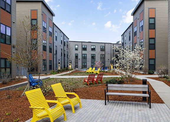 A playground area with a bench, chairs and a slide in front of apartment buildings.