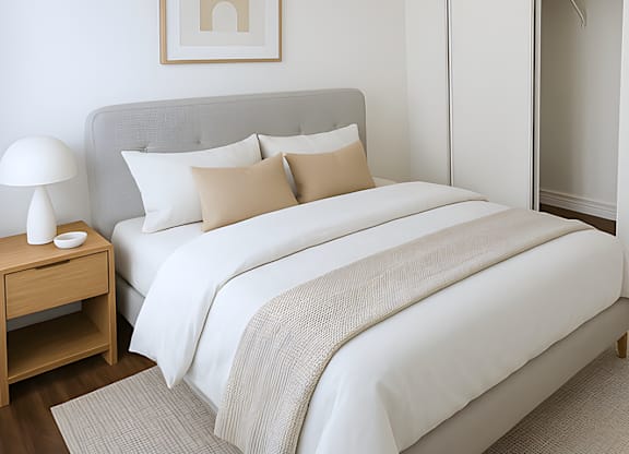 A bed with a grey headboard and white bedding is in a room with a wooden nightstand and a lamp.