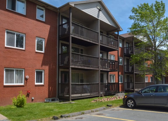 A grey car is parked in a parking lot in front of a red brick building with a balcony.