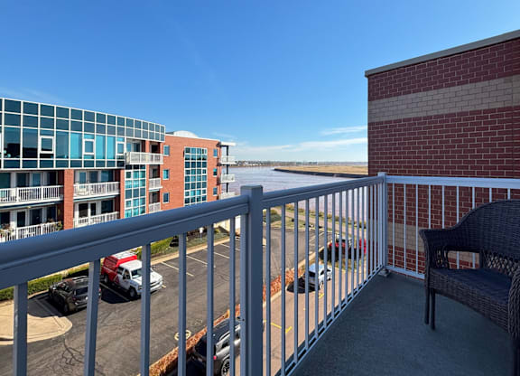 A balcony with a chair and a railing overlooking a parking lot and buildings.