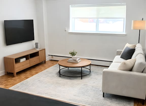 A living room with a grey couch, a wooden coffee table, a television, and a window with blinds.
