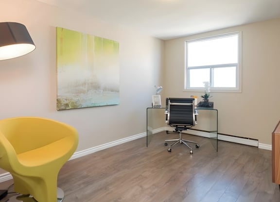 A yellow chair is in the corner of a room with a desk and a painting on the wall.