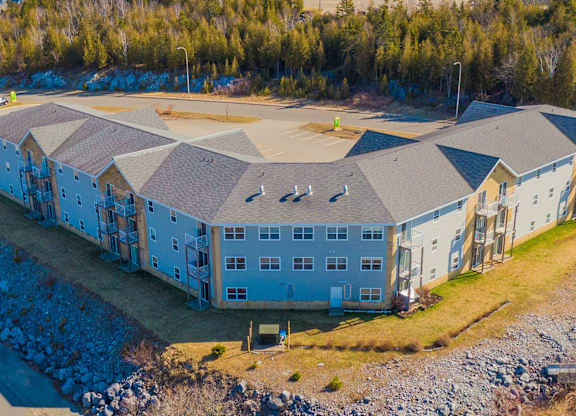 A large, multi-story building with a grey roof and white walls is surrounded by a rocky landscape and trees.