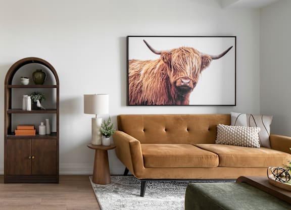 A living room with a tan couch, a brown side table, a lamp, a plant, a framed picture of a Highland cow, and a wooden cabinet.