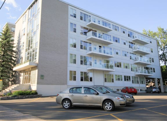 A silver car is parked in a parking lot in front of a white building with balconies.