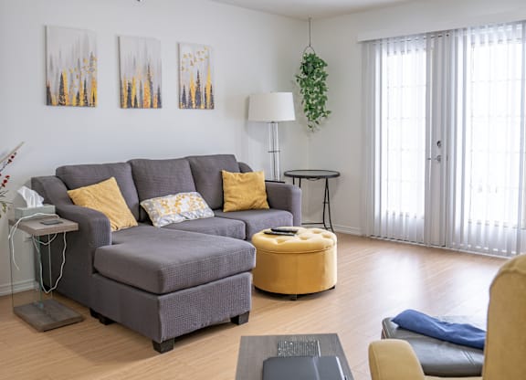 A living room with a grey couch, yellow ottoman, and a ceiling fan.