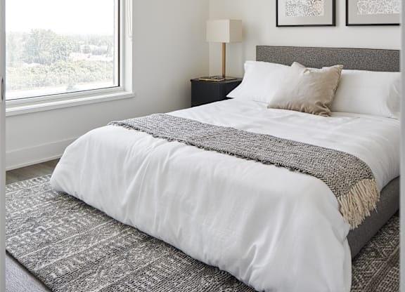A bed with a white comforter and a grey blanket is in the foreground with a lamp and two pictures on the wall.