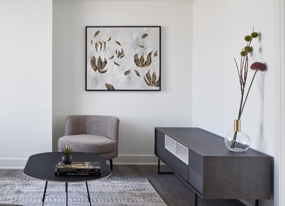 A grey chair sits in front of a painting and a coffee table with a vase on top.