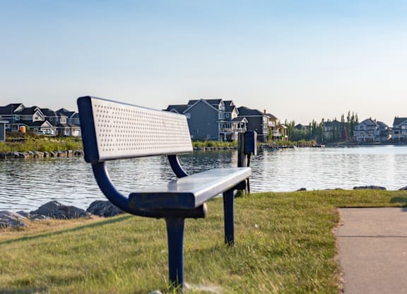 A bench is situated in front of a body of water with houses in the background.