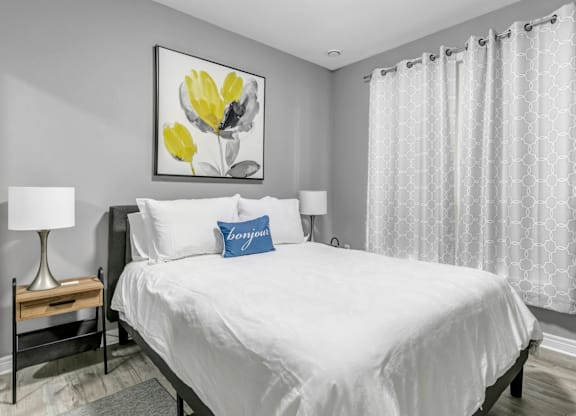 A bedroom with a large bed, a nightstand with a lamp, and a framed picture of yellow flowers on the wall.