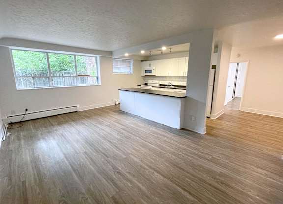 A spacious room with wooden flooring and a kitchen area in the background.