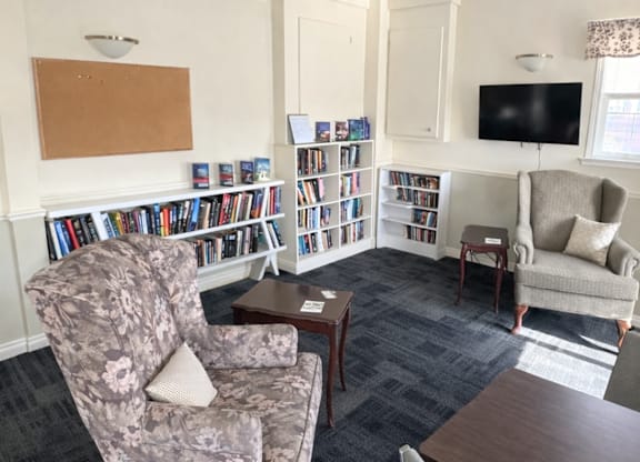 A living room with a grey floral armchair, a wooden coffee table, a grey sofa, a bookshelf with books, a television, and a window with curtains.