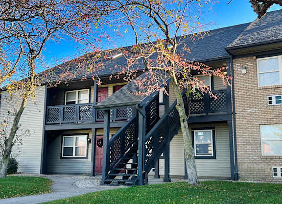 A tree with red leaves is in front of a grey house with a black staircase.