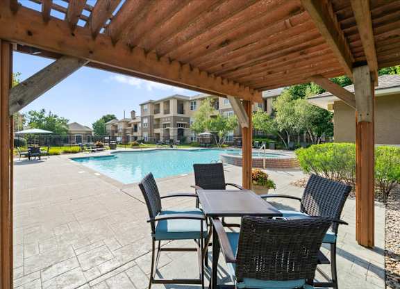 A wooden pergola over a table with chairs and a pool in the background. at Stonepost Lakeside Apartment Homes, Olathe, Kansas