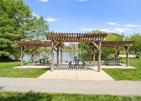 A wooden pergola with benches is situated in a park with a lake in the background at Stonepost Lakeside Apartment Homes, Kansas, 66062.