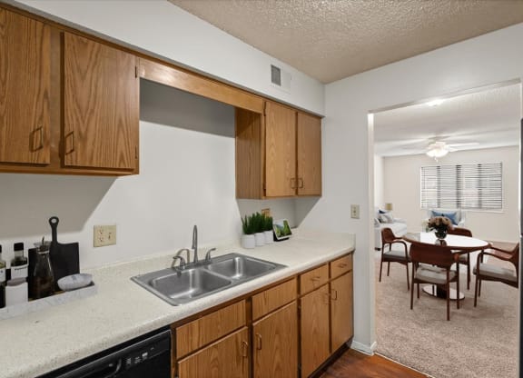 A kitchen with wooden cabinets, hardwood floors, white countertops, and a black dishwasher with a doorway to the dining area.