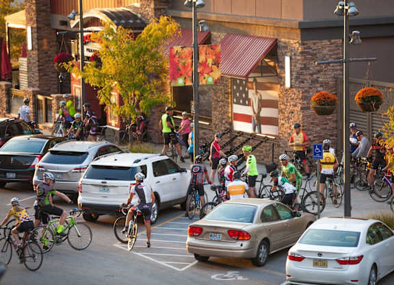 A group of cyclists are riding down a street in front of a building with a red awning. at Residences at Prairiefire Apartment Homes, Overland Park, 66223