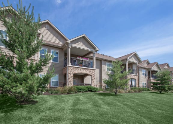 The exterior of an apartment building with large patios and balconies, overlooking a scenic grassy area with beautiful landscaping.