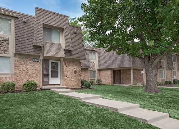 The exterior of an apartment building with a lush tree and walkway at Preston Court in Overland Park.