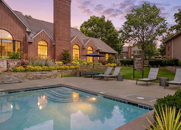 Twilight pool area with illuminated clubhouse windows, lounge seating, and landscaped grounds.