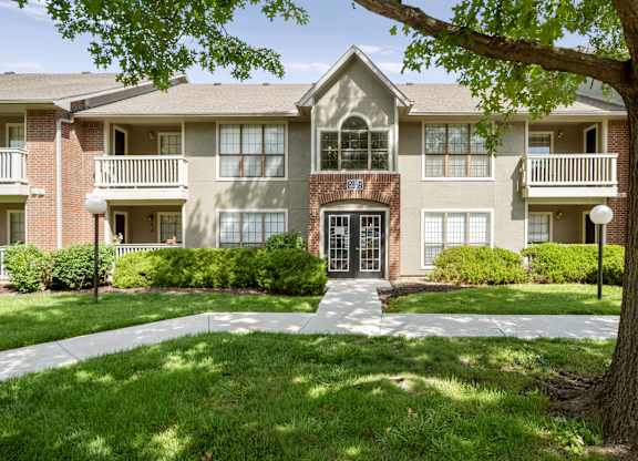 Front exterior of a residential apartment building with mature trees, walkways, and landscaping.