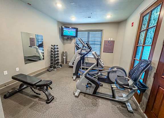 Compact fitness center at The Dorset Apartments featuring cardio machines, weight bench, dumbbells, and a mounted TV.
