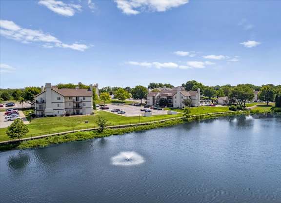 Decorative fountain in the community lake with apartment homes and mature trees nearby.
