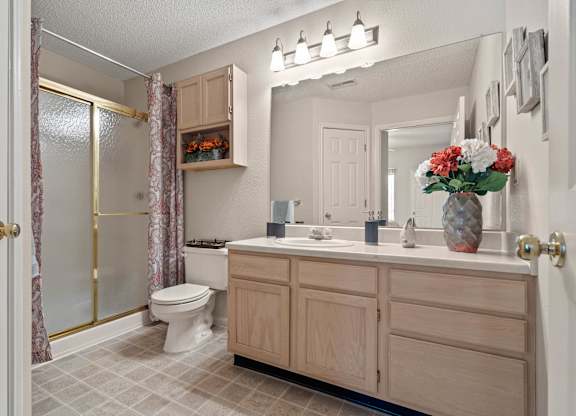 A bathroom with a sliding glass door shower, a linen closet, light wooden cabinets over the toilet, and a vanity with a large mirror and storage under the sink.