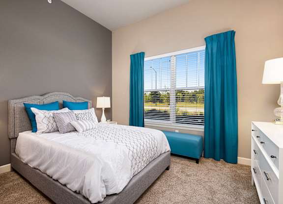 A bedroom with a large bed and a window with blue curtains. at Residences at Prairiefire Apartment Homes, Kansas, 66223