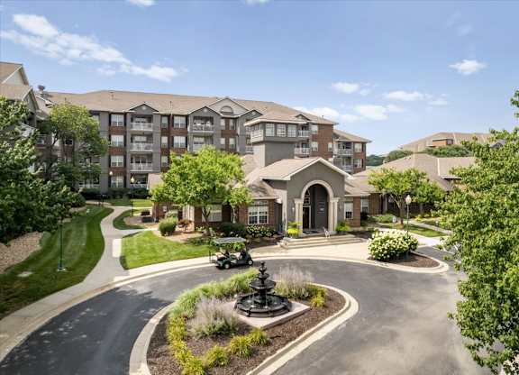 Wide aerial view of The Claremont Apartments entrance featuring a central fountain, mature trees, and clubhouse building in Overland Park, KS.