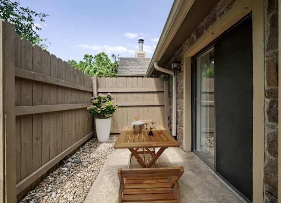 A wooden table and bench are on a patio with a stone wall and a potted plant.