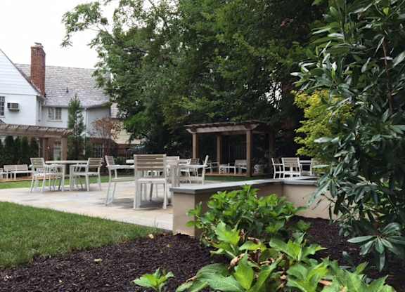 A patio with chairs and a table is surrounded by green plants.