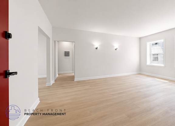 A spacious room with wooden flooring and white walls, featuring a red door and a window with a view of a building outside.
