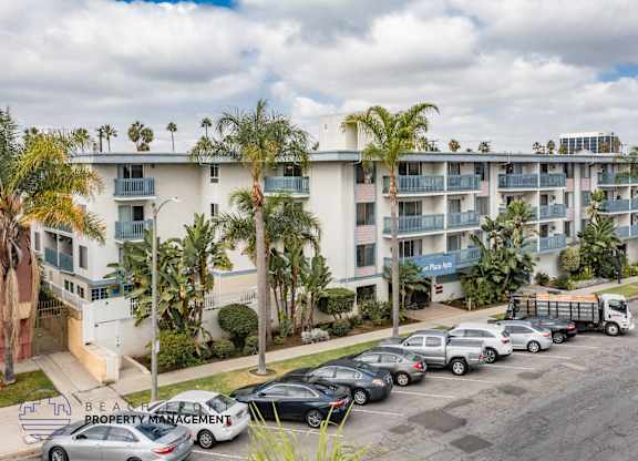 A parking lot with cars and a truck in front of a white apartment building with palm trees.