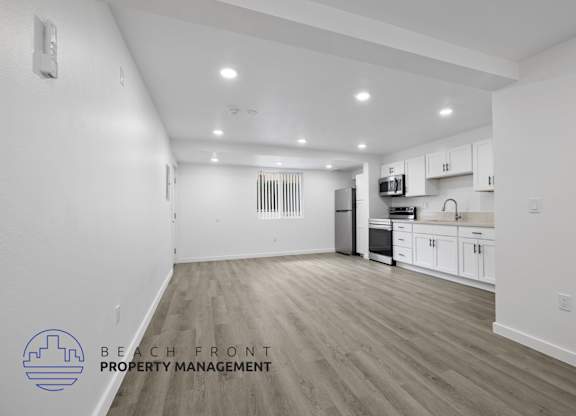 A spacious, empty room with a kitchen area in the background and a logo for Beach Front Property Management in the bottom left corner.