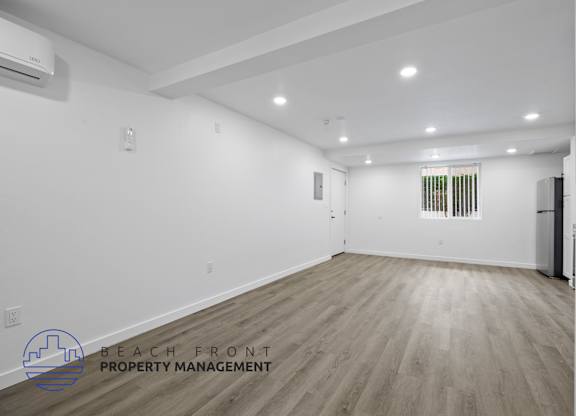 A spacious room with wooden flooring and white walls, with a logo for Beach Front Property Management in the bottom left corner.