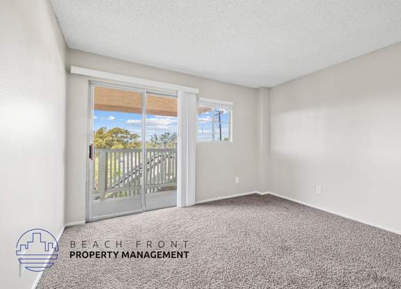 A room with a carpeted floor and a view of the beach through a sliding glass door.