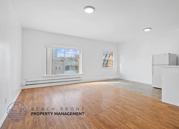 A spacious room with wooden flooring and a large window overlooking a beachfront.
