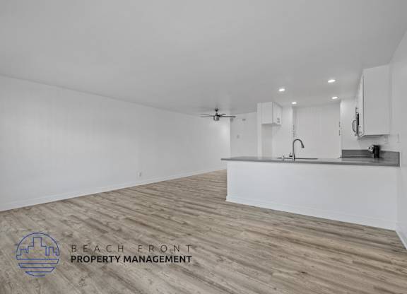 A spacious room with wooden flooring and white walls, featuring a kitchenette with a sink and a refrigerator.