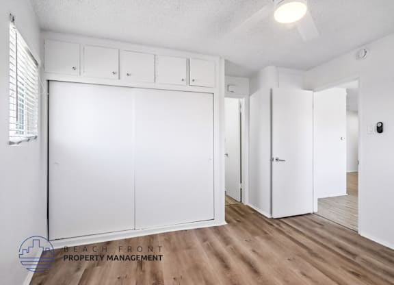A room with white walls and wooden flooring, featuring a row of white cupboards and a door on the right.